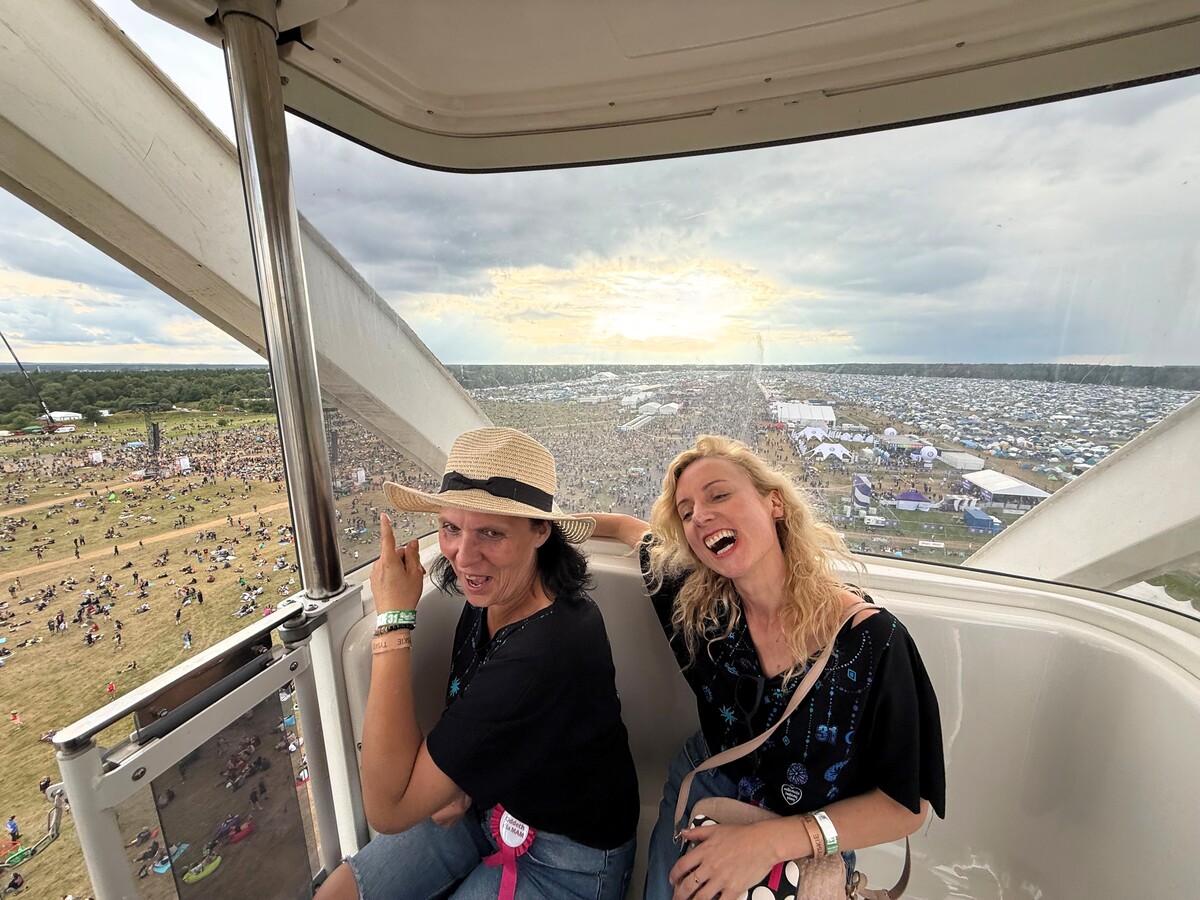 Two women sit in the gondola of the Ferris wheel, behind them the skyline of the festival with the crowd of people, the stage and the campground. One of the women wears a hat and points outside.